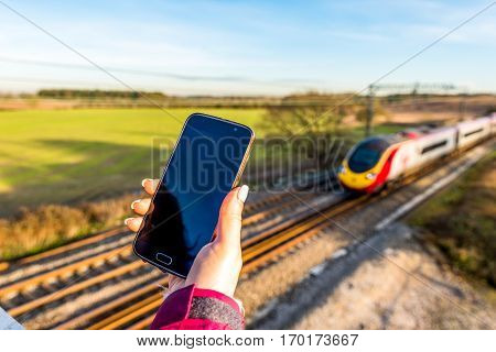 Day View Of Woman Hand Holding Smartphone Over Uk Railroad.