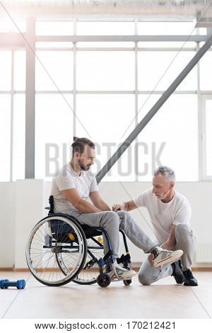Stretching the leg. Professional involved aged physical therapist helping the disabled man and providing a rehabilitation session while expressing concentration