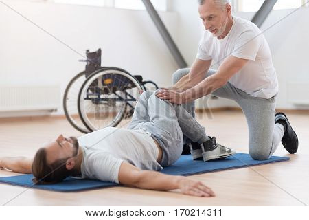 Working out the lower body. Attentive pleasant aged general practitioner stretching the handicapped and providing a rehabilitation session while holding legs of the patient