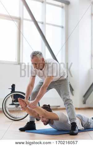 Working out with my disabled friend . Cheerful strong bearded orthopedist stretching the disabled and assisting while holding arms of the patient and expressing positivity