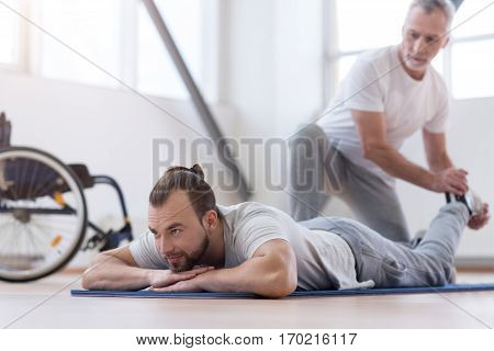 Helping my disabled son. Positive athletic bearded man stretching the disabled and assisting while holding legs of the young man and expressing positivity