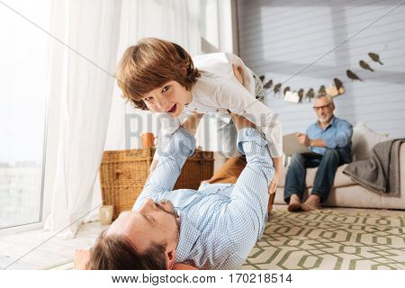 Lets fly. Delighted nice handsome father lying on the floor and holding his son while looking at him