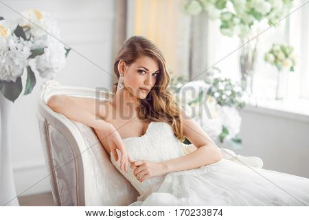 Wedding. Bride in beautiful dress sitting on sofa indoors in white studio interior like at home. Trendy wedding style shot. Young attractive caucasian brunette model like a bride against big window tender posing.