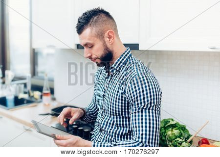 Cheerful Man Cooking In New Kitchen, Using Tablet For Receipes And Looking On Internet