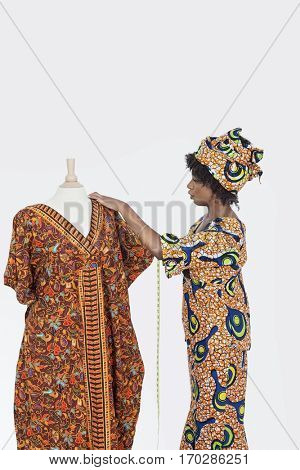 Female tailor in traditional outfit measuring dashiki on tailor's dummy over gray background