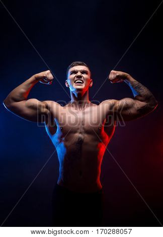 Handsome power athletic man bodybuilder. Fitness muscular body on dark smoke background. Perfect male. Awesome bodybuilder, tattoo, posing hands up. Viktory.