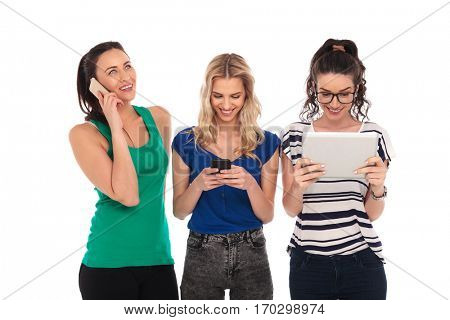 three young women having fun communicating. one looking up and talking on the phone, one sending an sms message and one reading on her tablet pad commputer on white backround