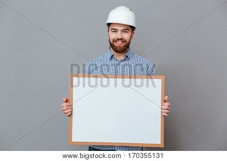 Image of attractve bearded builder holding copyspace board standing over grey background.