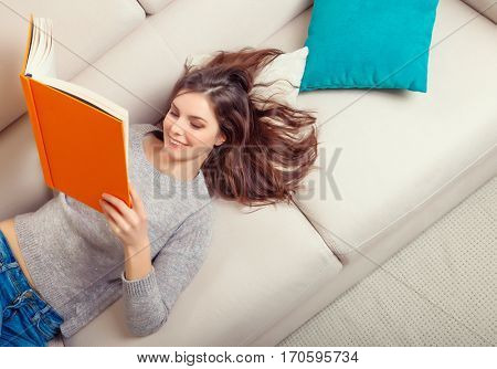Girl reads a book lying at white soft sofa. Culture lifestyle. Leisure. Top view. Young woman reading art literature