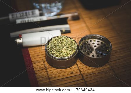 Steel or metal grinder full of freshly ground cannibus flower. Related smoking accessories nearby.