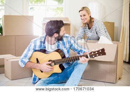 Man playing a guitar while woman unpackaging cardboard boxes in background in their new house