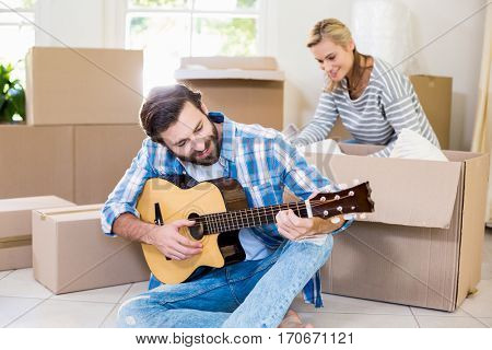 Man playing a guitar while woman unpackaging cardboard boxes in background in their new house