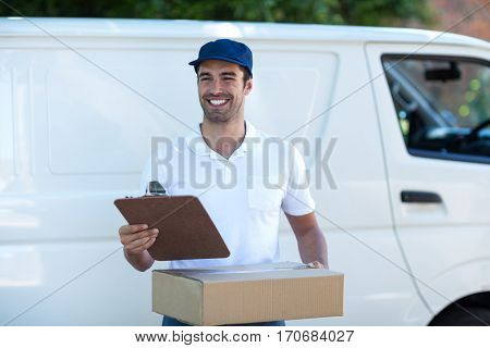 Smiling delivery man with clipboard and cardboard box while standing by van
