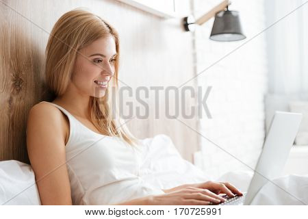 Side view of smiling woman lying on bed and using laptop in bedroom
