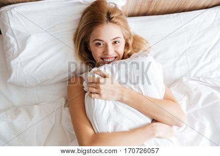 Top view of Happy woman lying on bed, embracing with pillow and looking at camera