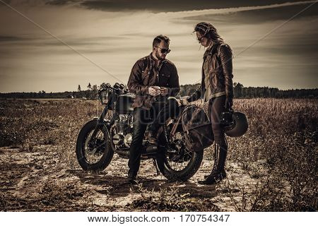 Young, stylish cafe racer couple on vintage custom motorcycles in field.