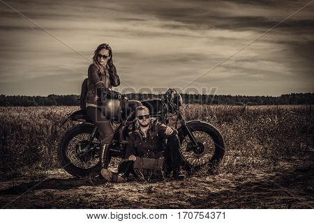 Young, stylish cafe racer couple on vintage custom motorcycles in field.
