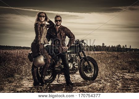Young, stylish cafe racer couple on vintage custom motorcycles in field.