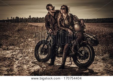 Young, stylish cafe racer couple on vintage custom motorcycles in field.