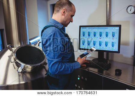 Side view of manufacturer holding digital tablet while working on computer at brewery