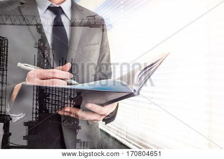 Double exposed of businessman writing some data in notebook with the building construction site.