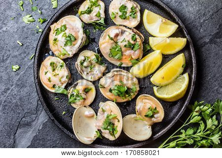 Fresh raw clams with lemon, herbs and white wine on black iron plate on stone slate background. Top view.