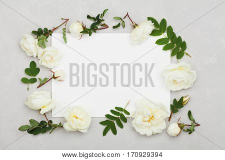 White rose flowers, green leaves and clean paper sheet on light gray background from above. Beautiful floral pattern, vintage color. Flat lay styling.