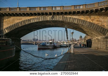 View of Prague castle under stone Palacky bridge, riverbank, Moldau river, Czech Republic, Europe, people walking on cobble stone pavement, anchored ships, white mute swans in water, sunny fall day