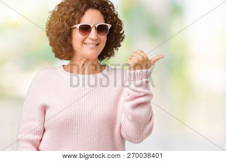 Beautiful middle ager senior woman wearing pink sweater and sunglasses over isolated background smiling with happy face looking and pointing to the side with thumb up.