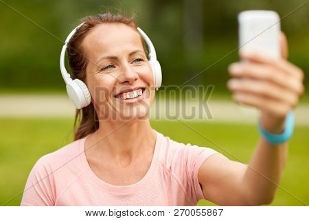 fitness, technology and healthy lifestyle concept - smiling woman in headphones listening to music and taking selfie by smartphone at summer park