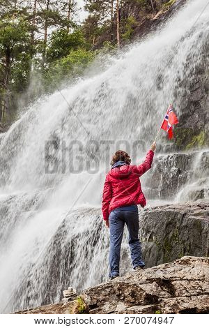Tourist Woman With Norwegian Flag At Svandalsfossen In Norway, Powerful Waterfall In Norwegian Mount