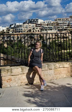 Young Blond Girl In Gray Dress With Pink Backpack Siting On The Stone Gate