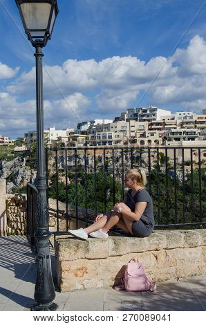 Young Blond Girl In Gray Dress With Pink Backpack Siting On The Stone Gate