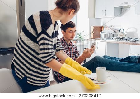 Lazy Man Looking At Phone While His Angry Girlfriend Cleaning His Dishes In Kitchen And Talking To H