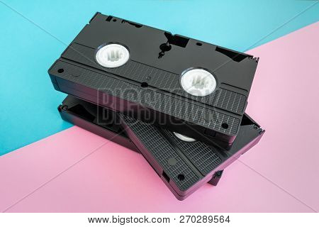 Stack Of 3 Black Vhs Video Tapes On A Bright Pink And Blue Background.