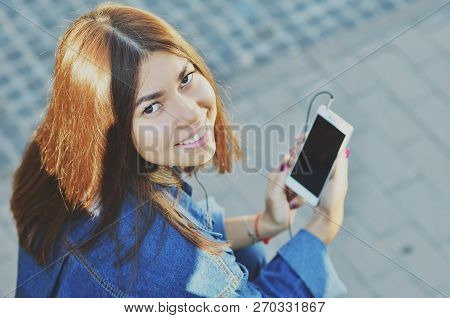 The Student Sits On A Bench And Listens To Music On The Phone With Headphones, Looks At The Camera S