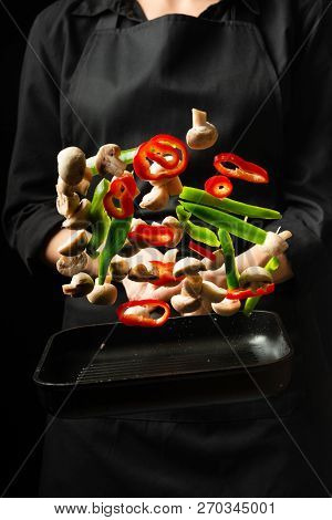 Professional Cook. The Chef Prepares A Dish With Vegetables And Mushrooms In A Saucepan. On A Black