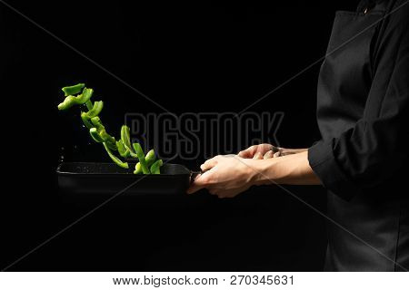 Professional Cook. He Prepares A Dish With Green Sweet Peppers In A Saucepan. On Black Background, M