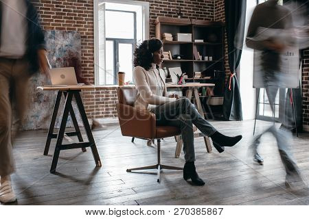 African American Businesswoman In Heaphones Sitting On Chair With Colleagues In Motion Blur In Moder