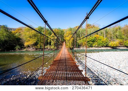 Narrow Metal Foot Bridge Across Mountain River In Autumn.