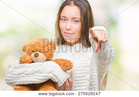 Young caucasian woman holding teddy bear over isolated background pointing with finger to the camera and to you, hand sign, positive and confident gesture from the front