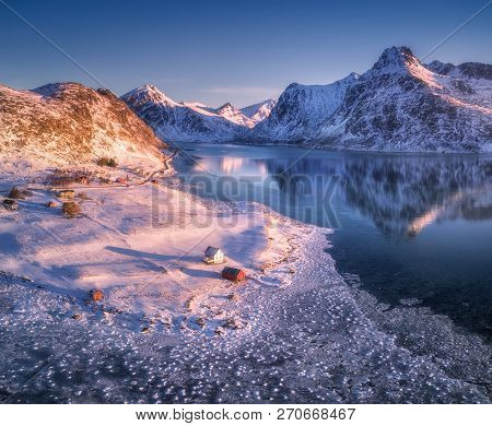 Aerial View Of Snow Covered Mountains, Houses, Frosty Seacoast And Blue Sky Reflected In Water At Su