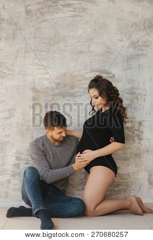 Happy Man Put His Handr On The Belly Of His Pregnant Wife. Young Family In Studio. Couple Wait For A
