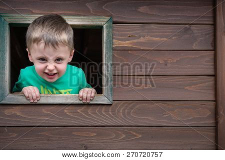 Portrait Of A Cute Cheeky Little Caucasian Boy Looking Through The Window Of A Wooden Toy House In A