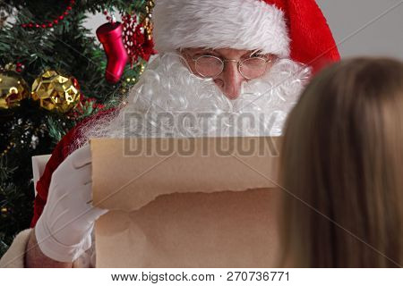 Santa Claus With With List Or Nice Or Naugthy List Paper With Small Boy Near Christmas Tree