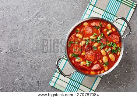 Hearty Beans Stew With Sausages, Herbs And Spices In Tomato Sauce In A Metal Casserole On A Concrete