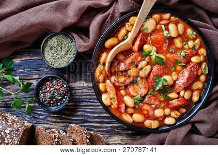 Overhead View Of Hearty Beans Stew With Sausages, Herbs And Spices In Tomato Sauce In A Black Bowl W