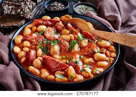 Beans Stew With Sausages, Herbs And Spices In Tomato Sauce In A Black Bowl With Spoon, On A Rustic W