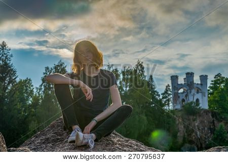 Pretty Young Lady With Red Hair Sitting On Stone Near Bay Shore In Summer Evening. Tourist On The Be