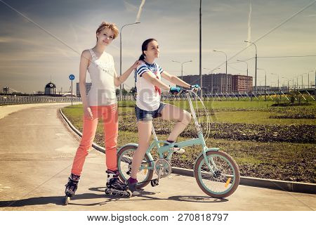 Two Young Female Friends One On A Bicycle, One On A Roler Scates. Summer Sport And Leisure Theme. Wa
